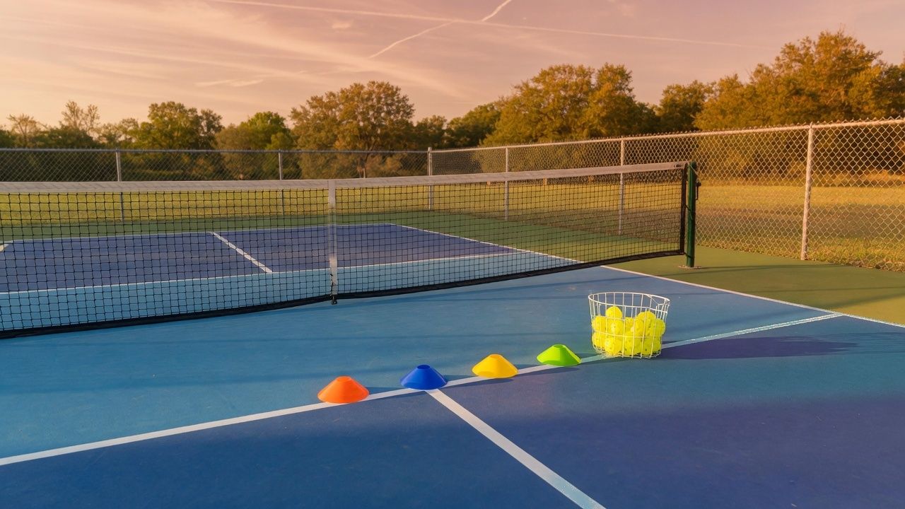 Empty blue-and-green pickleball court at golden hour, with four colored cones on the kitchen line, a wire basket of perforated balls, and one low net
