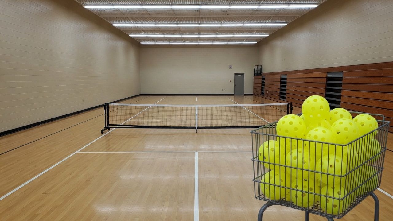 Empty community-center gym with pickleball lines on a hardwood floor, one portable net across the court, and a cart of perforated balls