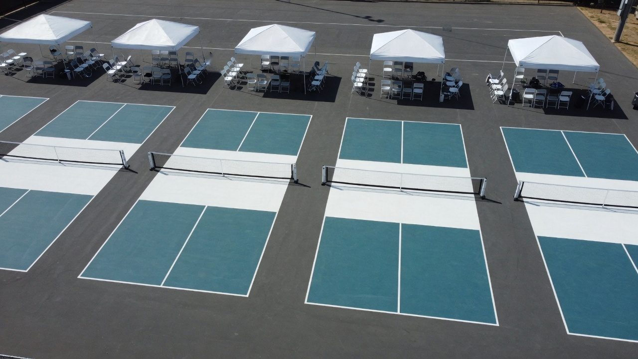 Elevated view of several empty outdoor pickleball courts in a row, each with one low net, white canopy tents, and empty folding chairs